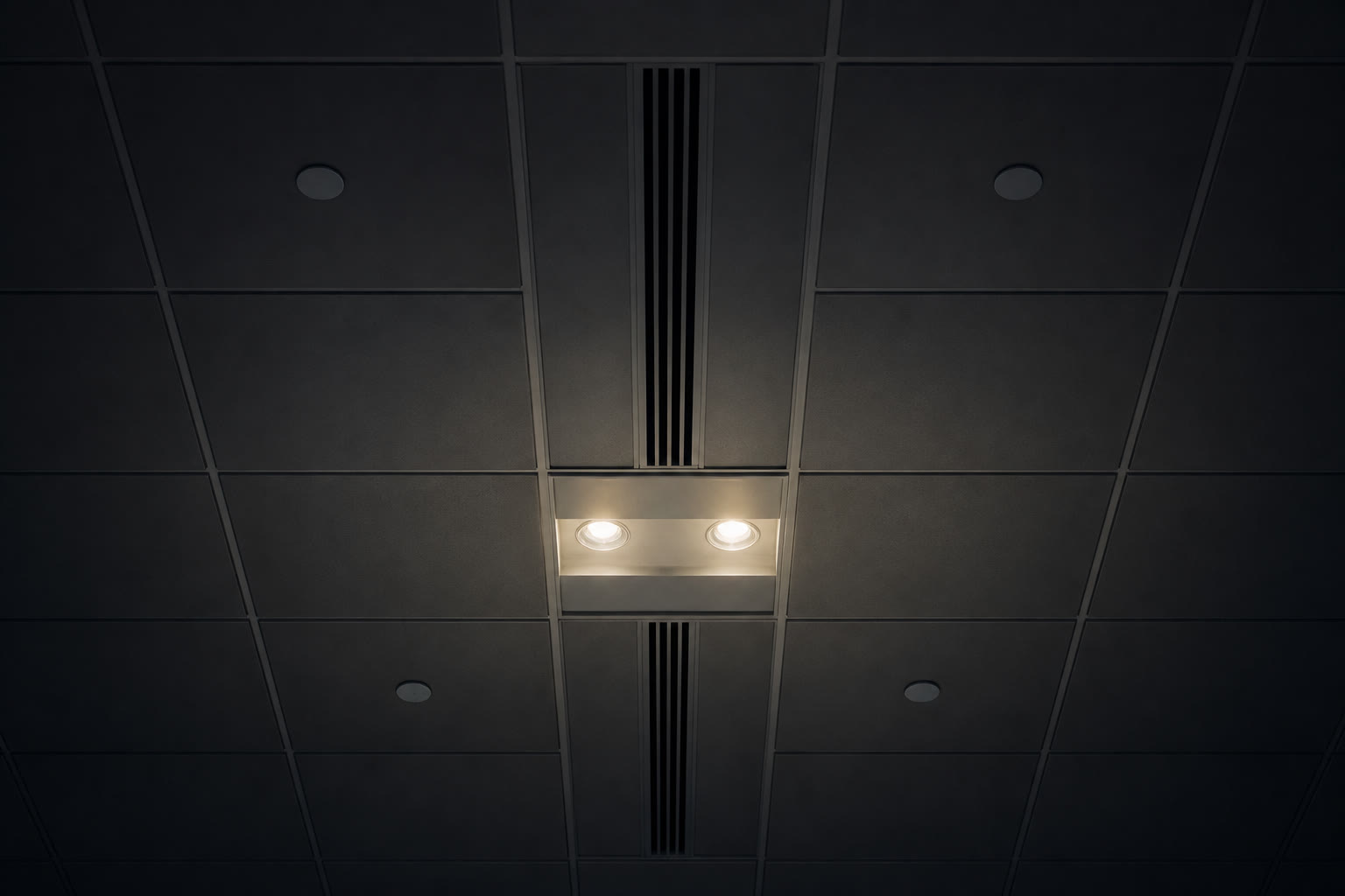An office ceiling seen from directly below at night, a grid of tiles with small flush-mounted sensor discs, linear diffusers and downlights, one bay lit and the rest dark.