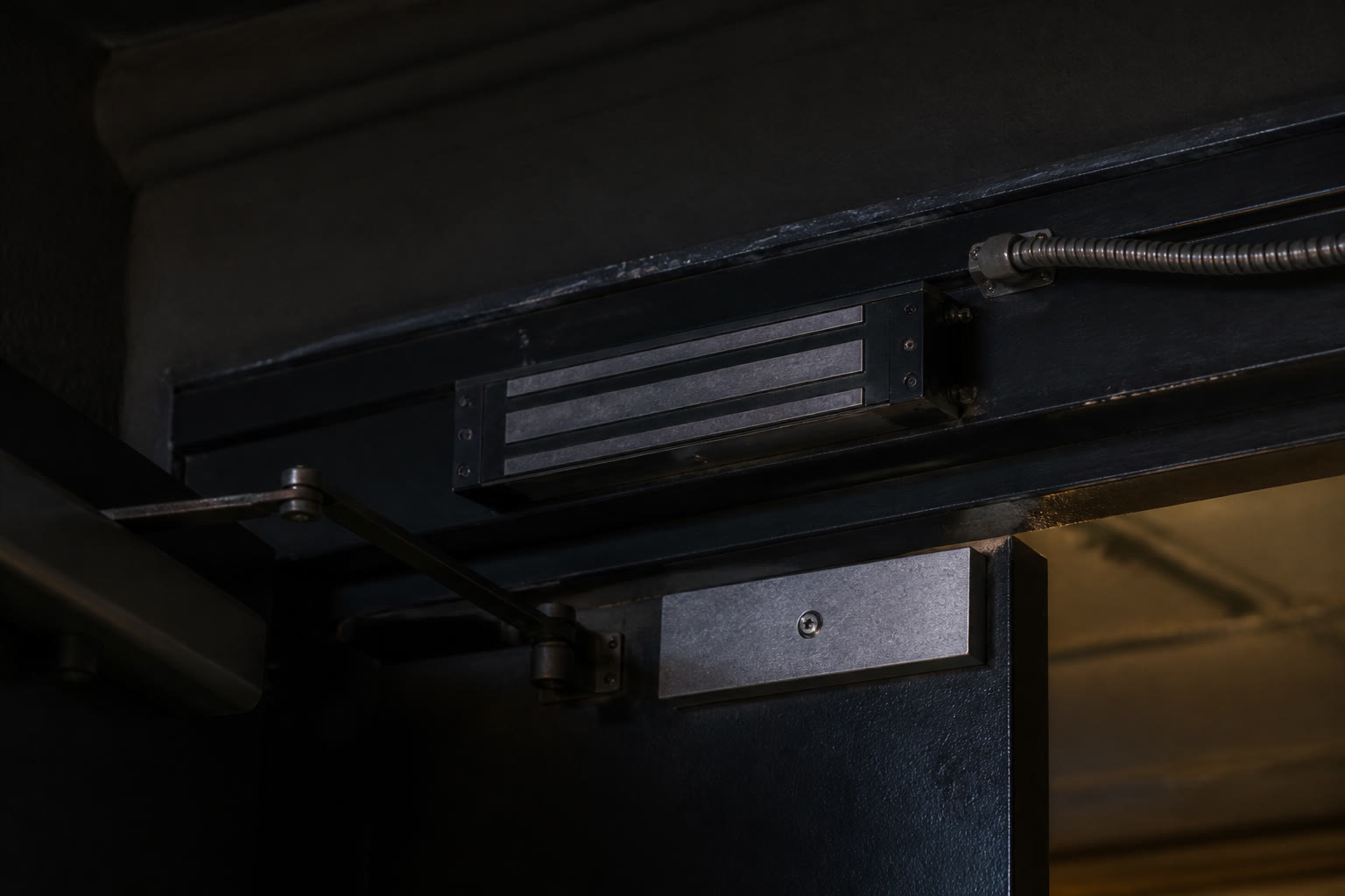 A close view of the top corner of a heavy commercial door at night, an electromagnetic lock and armature plate on the frame with the door closer arm above.