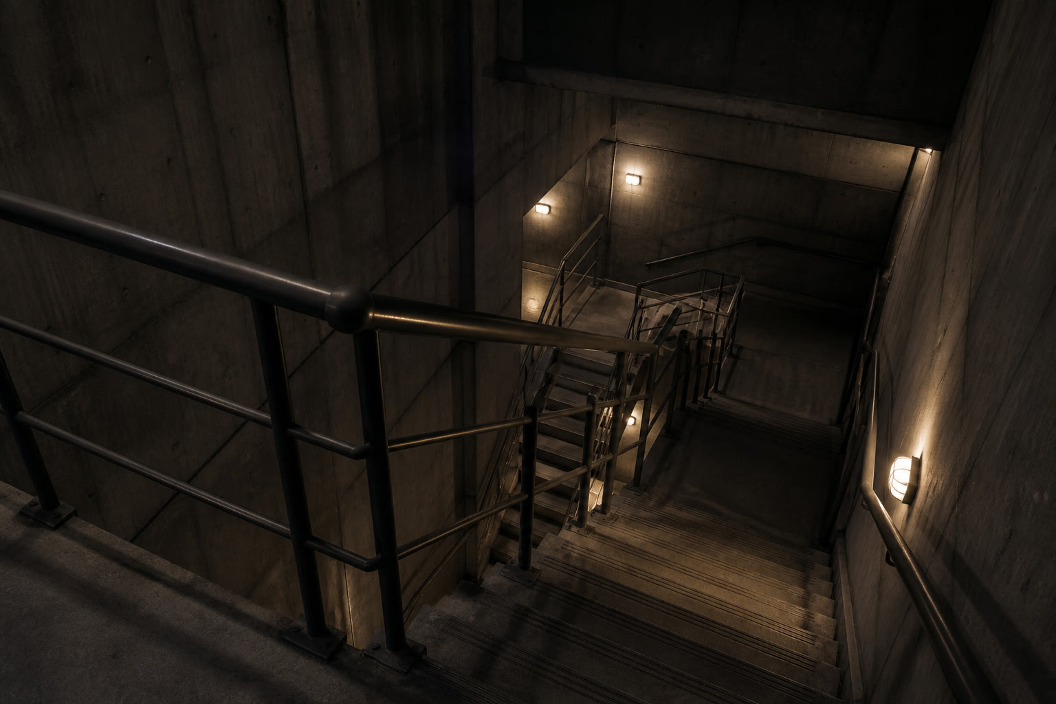 A concrete escape stair core at night lit by bulkhead lights, steel handrails switching back down several flights.