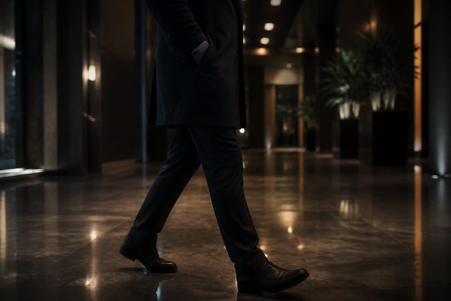 A person seen from the waist down walking through a dim lobby at night, a smartphone half out of a coat pocket, the polished floor reflecting overhead light.