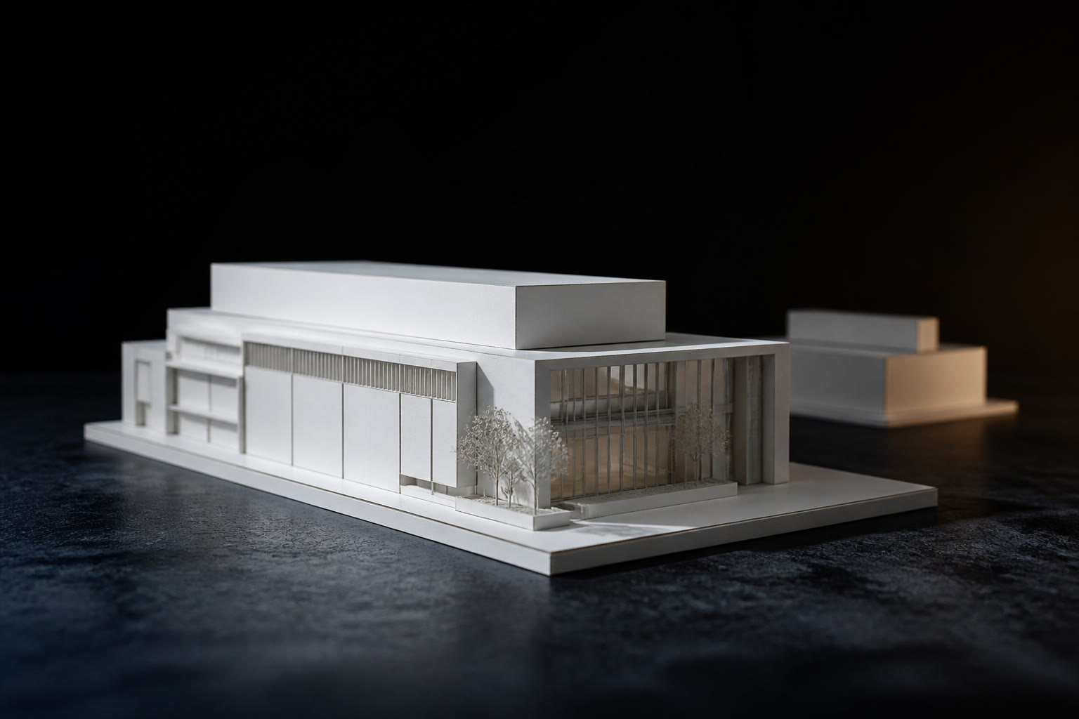 A white architectural scale model of a commercial building on a dark table, lit from one low side so the massing throws long shadows.